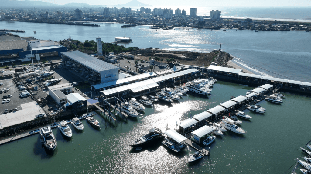 Vista aérea da Marina de Itajaí com diversos barcos e iates atracados nos píeres, galpões cobertos ao centro e a cidade ao fundo, refletidos nas águas sob a luz do sol.