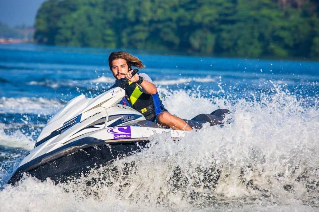 Homem usando colete salva-vidas pilotando um jet ski branco e roxo em alta velocidade, levantando grande borrifo de água.