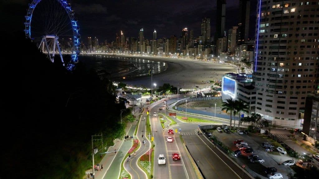 Vista noturna da orla de Balneário Camboriú, com a roda-gigante Unipraias iluminada em azul e as avenidas e prédios acesos