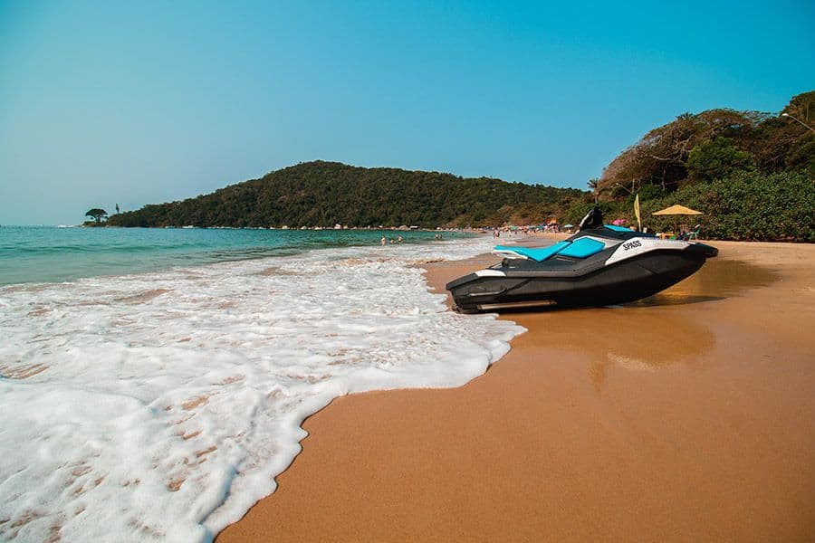 Jet ski estacionado na faixa de areia da Praia de Laranjeiras, com espuma suave das ondas na beira do mar e uma colina arborizada ao fundo.