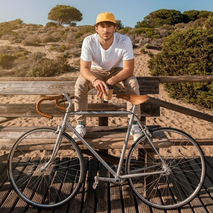 Homem sentado em banco de madeira em passarela de praia segurando um copo de café, com bicicleta apoiada à frente e vegetação ao fundo.