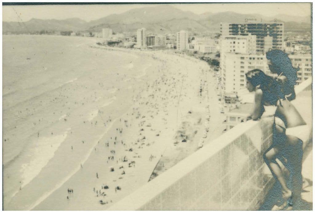 Imagem em preto e branco de duas mulheres de biquíni apoiadas em uma varanda, observando a praia e os prédios do litoral.