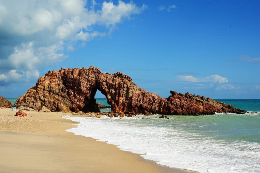 Pedra Furada em Jericoacoara (CE): arco de rocha avermelhada sobre faixa de areia clara e águas turquesa, com céu azul ao fundo